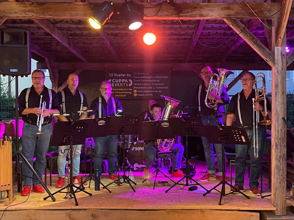 Die Combo auf einer Bühne unter Holzdach, pinkes und violettes Bühnenlicht, alle stehen mit Instrumenten.