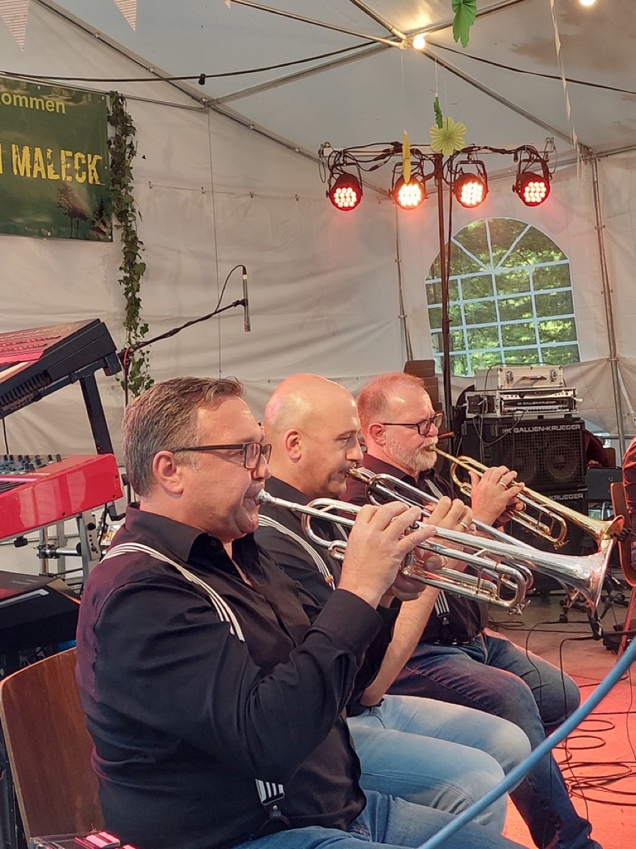 Drei Trompeter sitzen nebeneinander beim Auftritt im Festzelt, schwarzer Look mit Hosenträgern.
