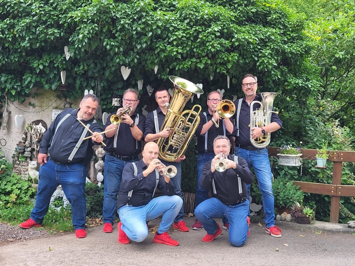 Die sieben Musiker der Combo blech'n'takt mit Trompeten, Posaune, Tuba, Euphonium und Schlagzeug-Sticks vor grüner Hecke. Schwarzer Look mit Hosenträgern und roten Schuhen.